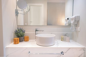 A bathroom with a round white sink, a mirror, a hairdryer, and a potted lavender plant.