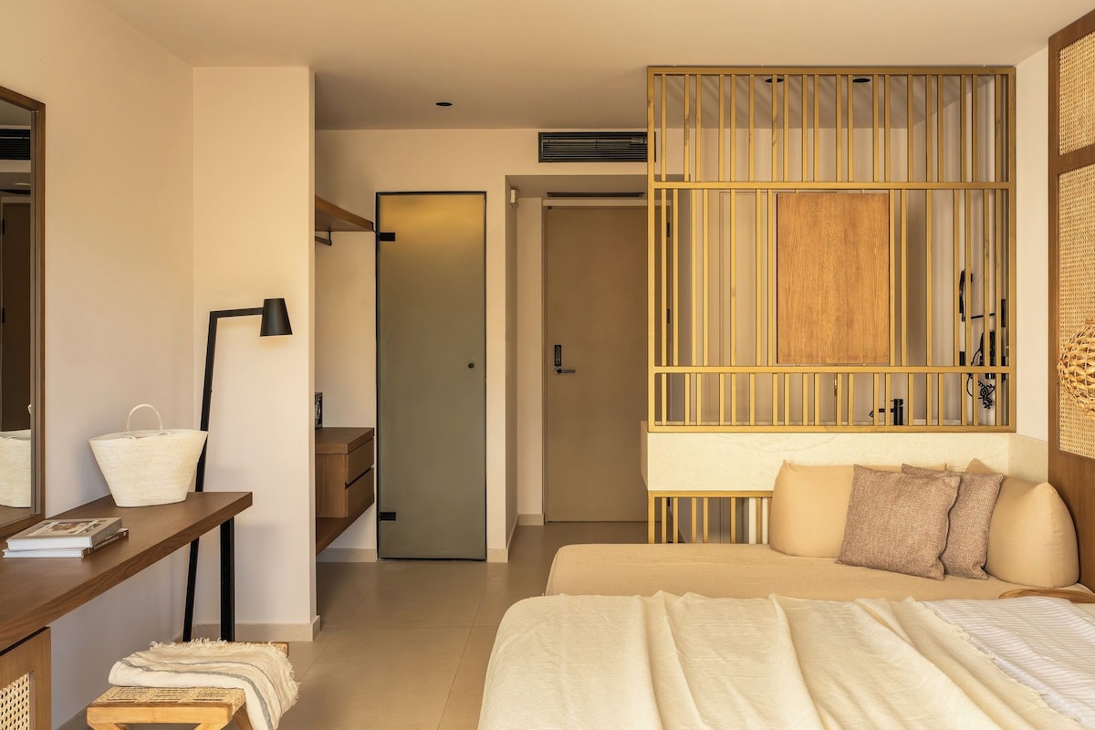 Habitación de hotel moderna con cama, mesita de noche, lámpara y vista al pasillo.