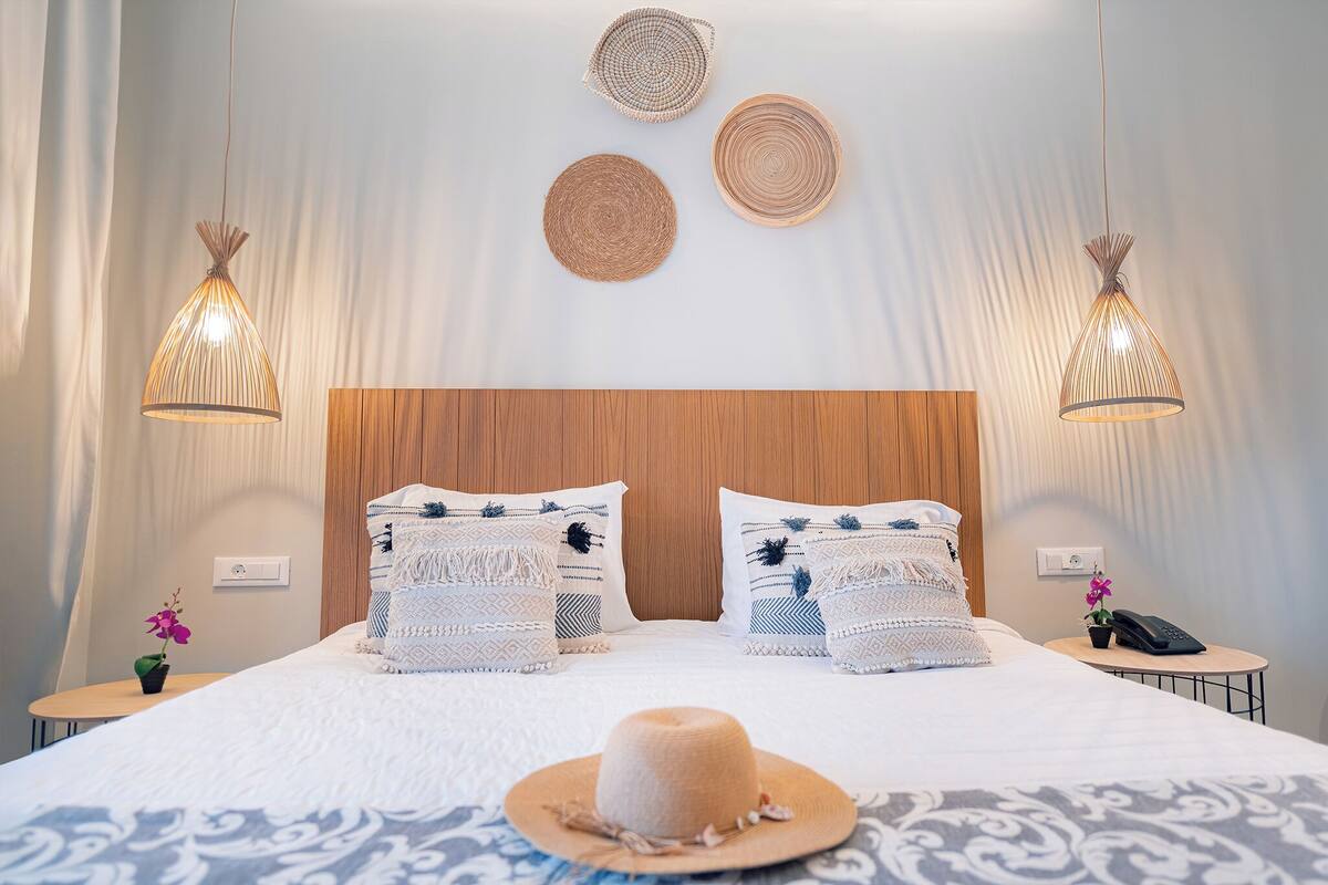 A neatly arranged hotel room with a large bed, two bedside tables, a wooden headboard, and two hanging light fixtures.