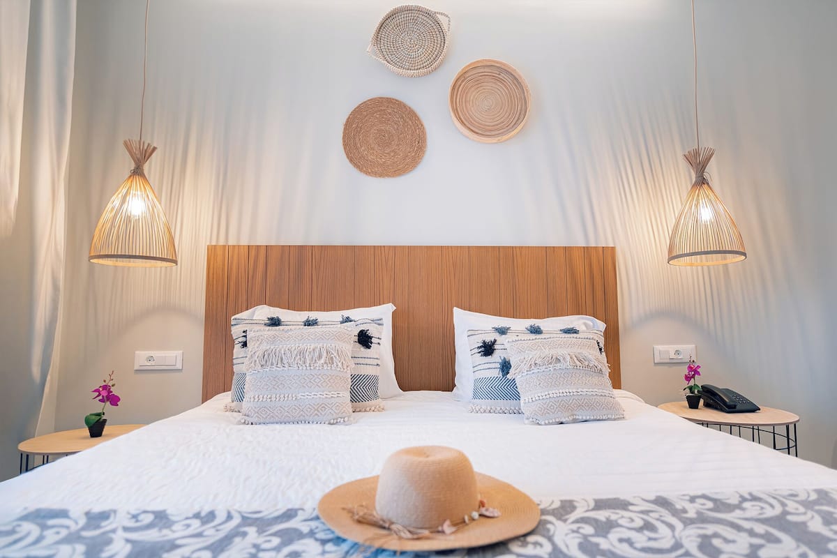 Habitación de hotel ordenada con una cama grande, dos mesitas de noche, un cabecero de madera y dos lámparas colgantes.