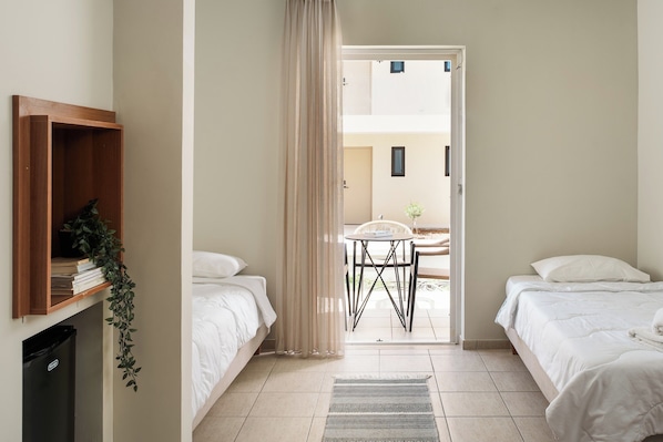A hotel room with two beds, a wooden shelf with books and a plant, a small table with a chair, and a view of the outside.