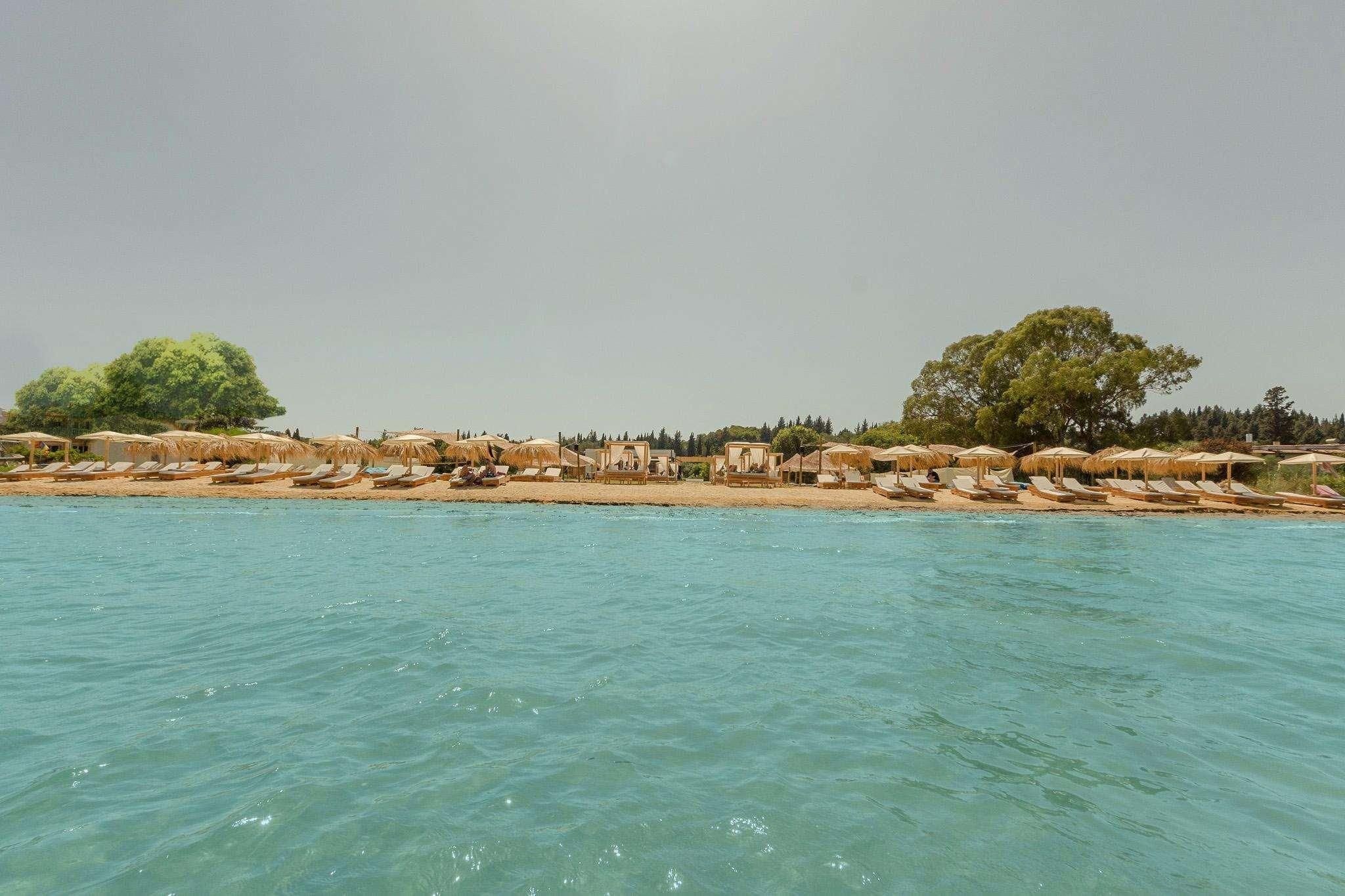 Ein Hotel am Strand mit Reihen von Liegestühlen und Reetdachschirmen, einem klaren, blauen Meer und Bäumen im Hintergrund.