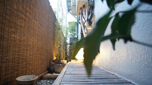 Un étroit sentier mène à l’entrée d’un bâtiment, avec un mur de bambou d’un côté et un mur blanc de l’autre.