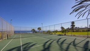 En utomhusbana för tennis med fri utsikt över havet, omgiven av palmer och ett staket.