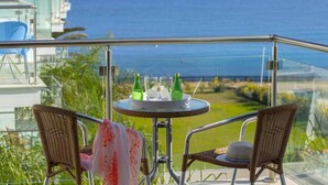 En balkong med ett glasräcke, två flätade fåtöljer, ett litet runt bord med ett bricka fullt av gröna flaskor och glas, och utsikt över havet.