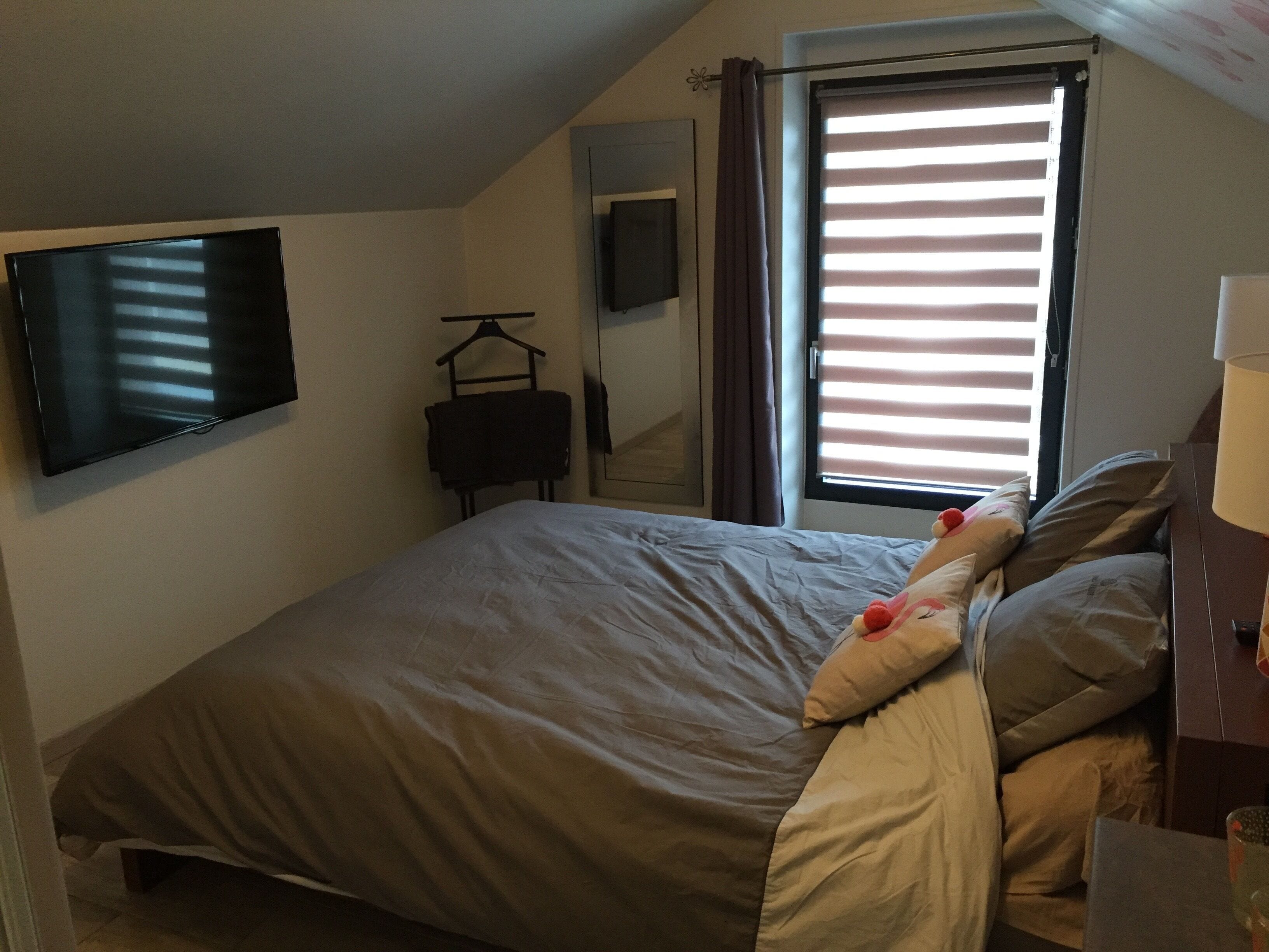 Un dormitorio con una cama, un televisor montado en la pared, una ventana con persianas y una lámpara sobre una mesita de noche.