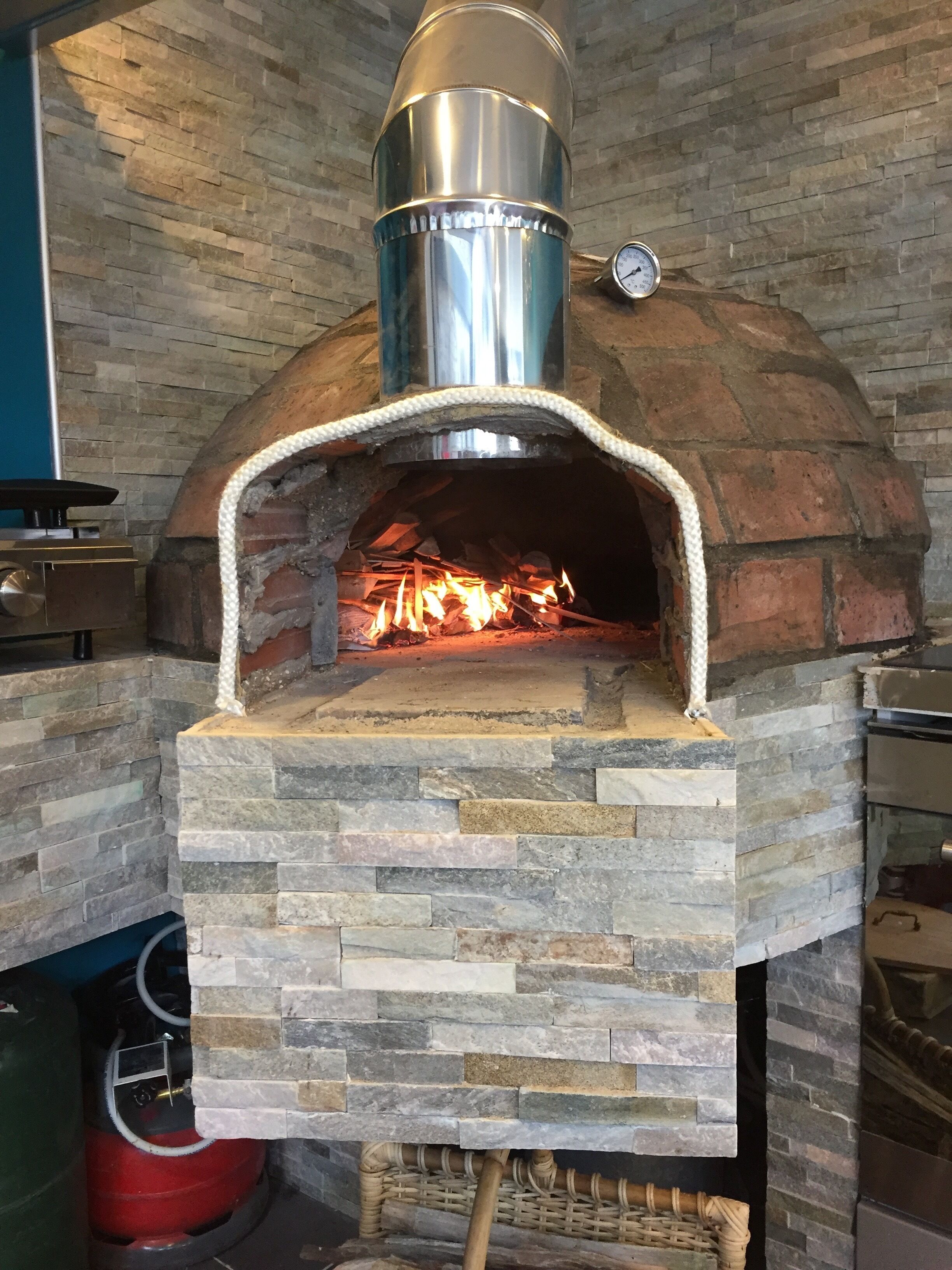 A brick oven pizza oven with a chimney, stone walls, and a fire burning inside.