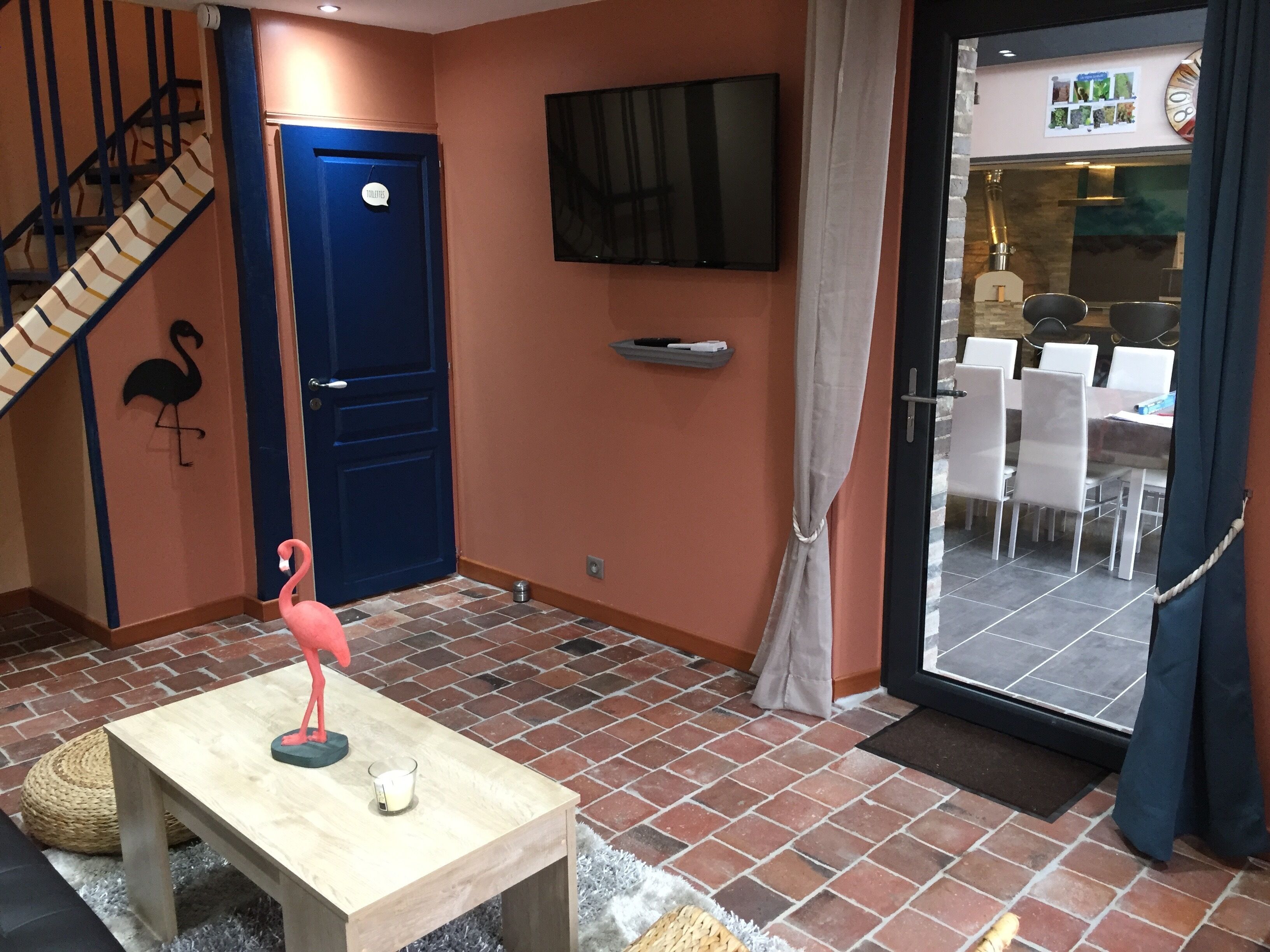 Une pièce avec une table en bois, un objet décoratif en forme de flamant rose, un téléviseur fixé au mur et un escalier avec une porte bleue portant l’inscription « TOILETTES ».