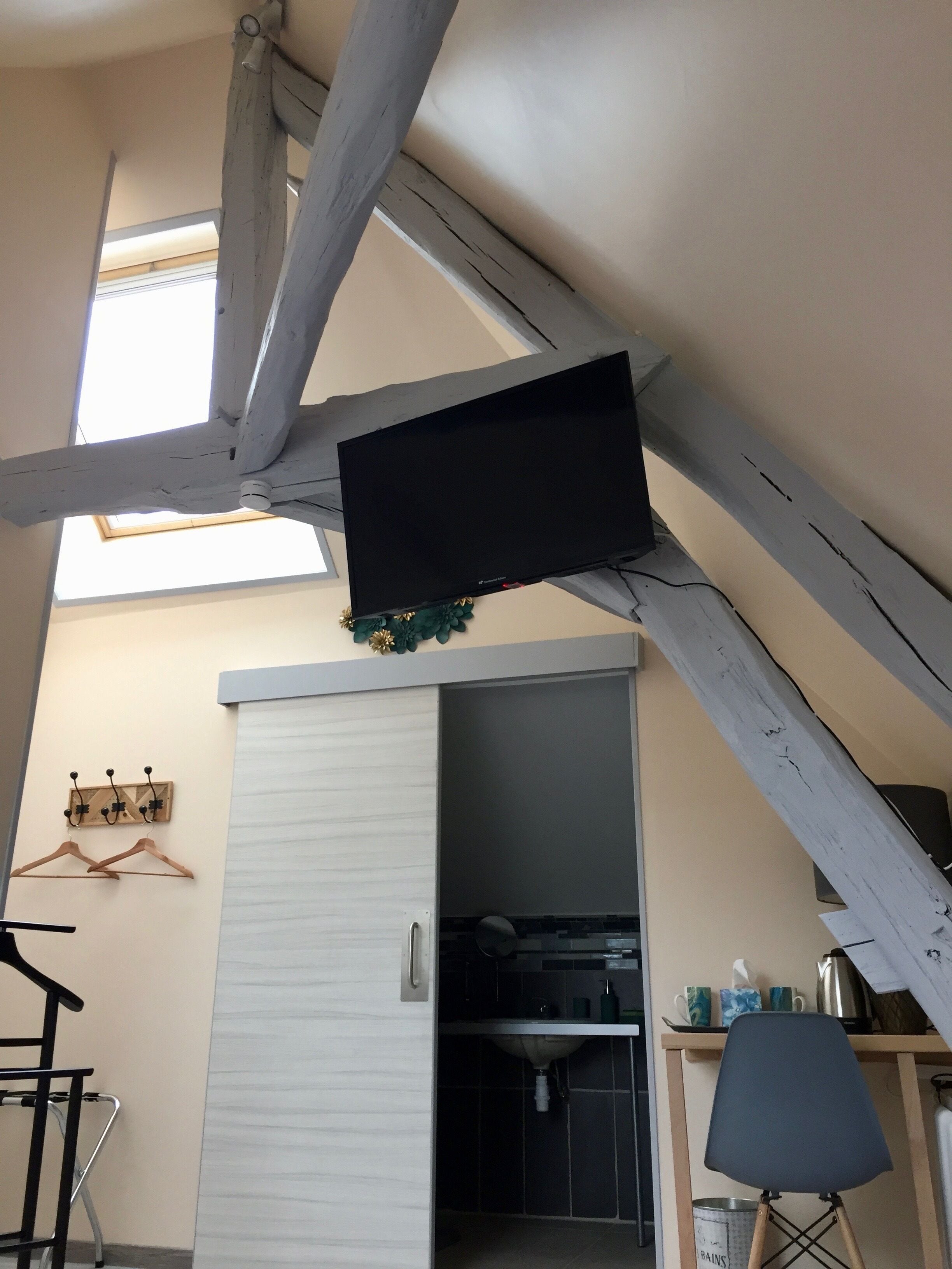 A room with a sloped ceiling, a black flat-screen TV mounted on the ceiling, a wooden chair, a small table, and a kitchen area with a sink and cabinets.