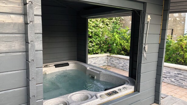 A hot tub with a stone exterior, a wooden deck, and a view of greenery. - Le Rouge & Blanc (Saint-Maurice-le-Vieil)