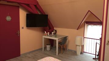 A small, cozy room with a red door, a flat-screen TV mounted on the wall, a small table with a chair, a bedside lamp, and a window with a view.