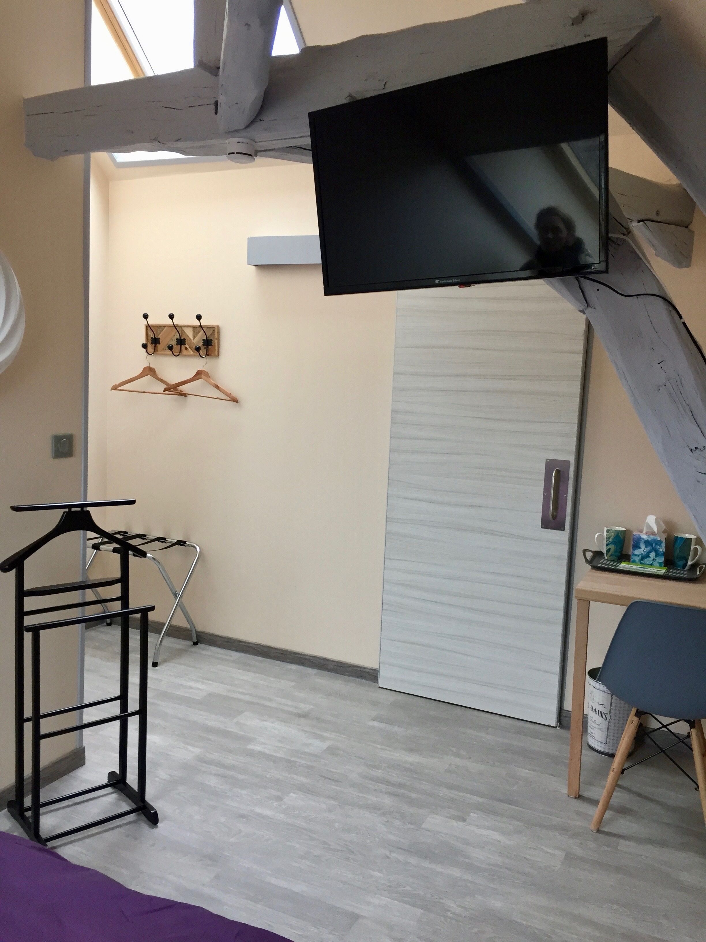 A room with a TV mounted on the wall, a coat rack, a folding chair, and a small table with cups and a tissue box.