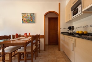A dining area with a wooden table and chairs, a painting on the wall, and a kitchen with a microwave and lemons on the counter.
