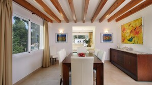 A dining area with a wooden ceiling, white walls, and a large window with curtains. There are two paintings on the walls, a dining table with chairs, and a kitchen area with a wooden cabinet. - MEDINA. Modern villa just 325 m from crystal clear beaches (Cala San Vicente)