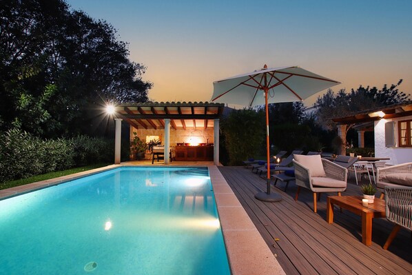 Una piscina con muelle de madera, sillas de descanso y una pérgola con mesa y sillas debajo.