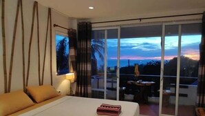 Une chambre d’hôtel avec un lit, une fenêtre donnant sur le coucher de soleil et un balcon avec une table et des chaises.