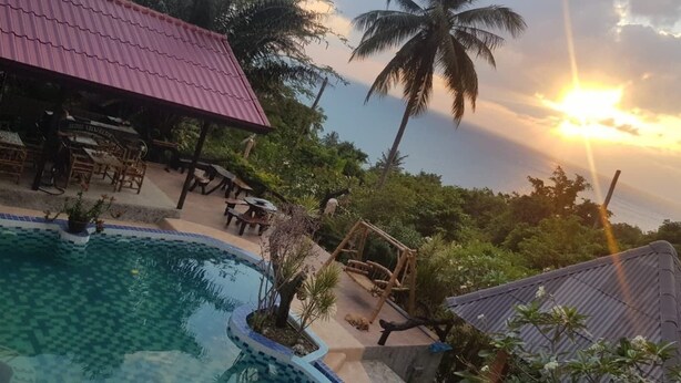 Un espace piscine avec un bâtiment au toit rouge, des palmiers et un coucher de soleil.