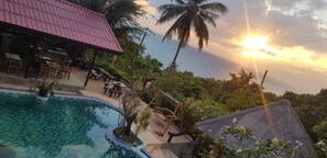 A pool area with a red-roofed building, palm trees, and a sunset. - Sairee View Resort (Koh Tao)