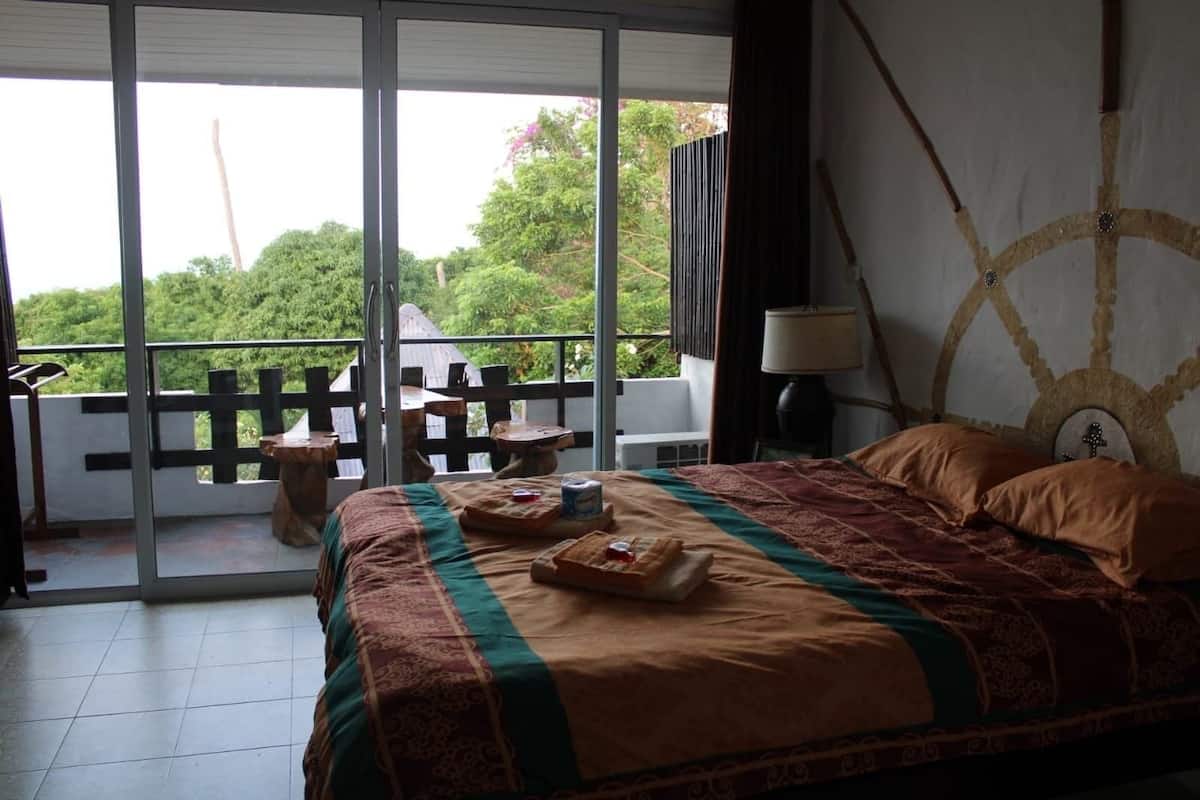 Ein Schlafzimmer mit einem großen Bett, Blick ins Grüne durch eine Schiebetür und eine Holzterrasse vor der Tür.