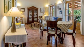 Un comedor con una mesa lista para cuatro personas, sillas de madera, un armario con vajilla y una lámpara sobre una mesita.