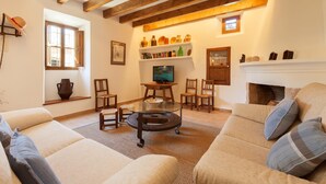 Een gezellige woonkamer met een bank, stoelen, een glazen salontafel, houten balken, een open haard en planken met decoratieve accessoires.