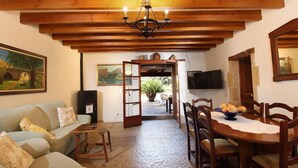 A cozy living room with a wooden ceiling, a dining table with chairs, a sofa, and a TV mounted on the wall.