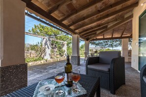 An outdoor patio with wicker furniture, a table with wine glasses and a bottle, and a view of greenery and buildings in the background.