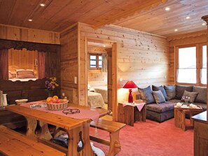 A cozy log cabin interior with a wooden dining table, benches, a sofa, and a small table with a lamp.