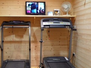 Una habitación con dos cintas de correr, un televisor sobre una estantería de madera y un ventilador.