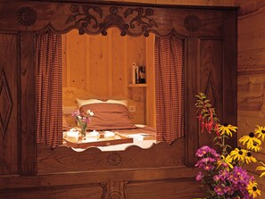 Una habitación de madera con una cama, una mesita con un florero y una estantería con libros.