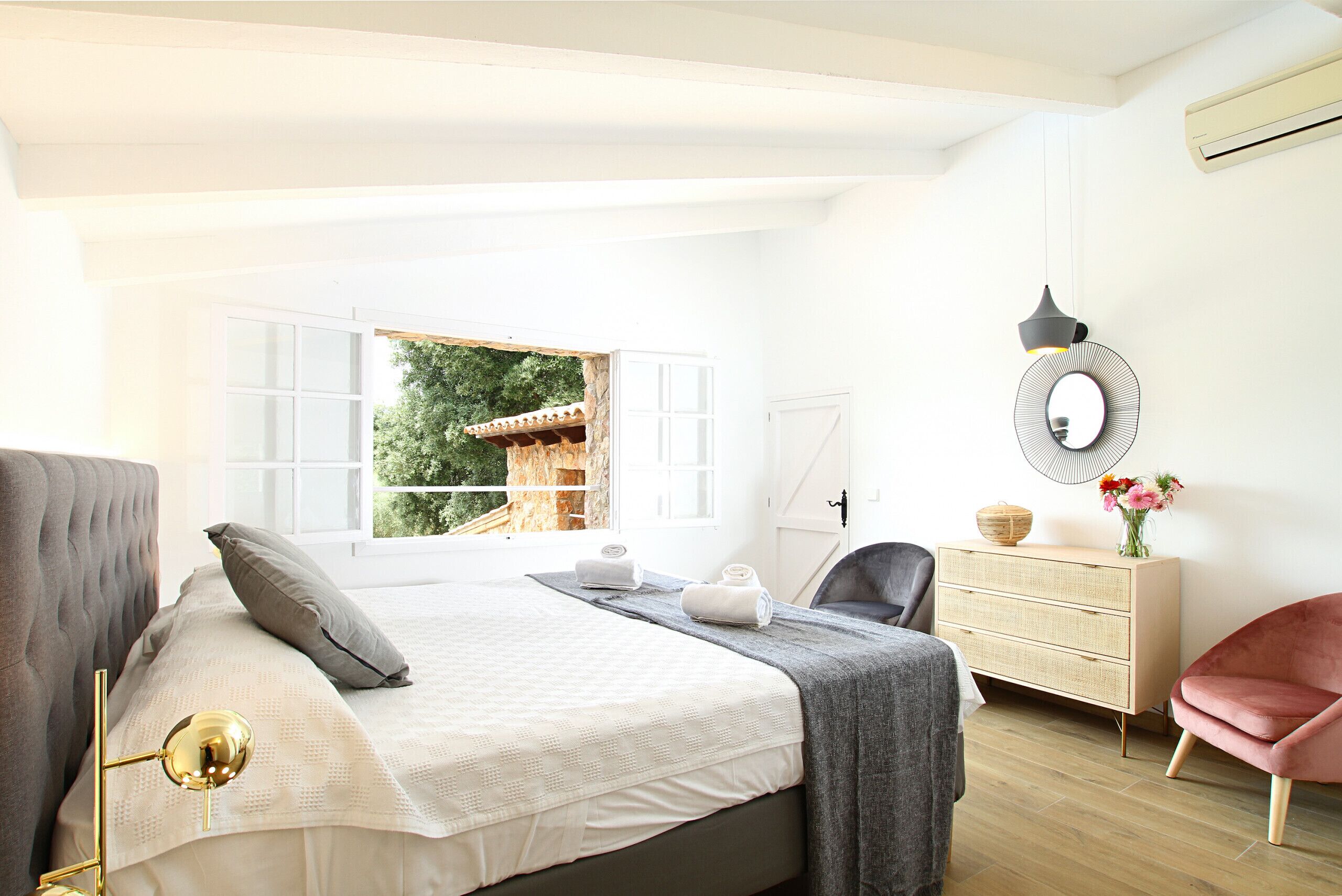 A bedroom with a bed, bedside lamp, dresser, chair, and a window with a view of greenery.