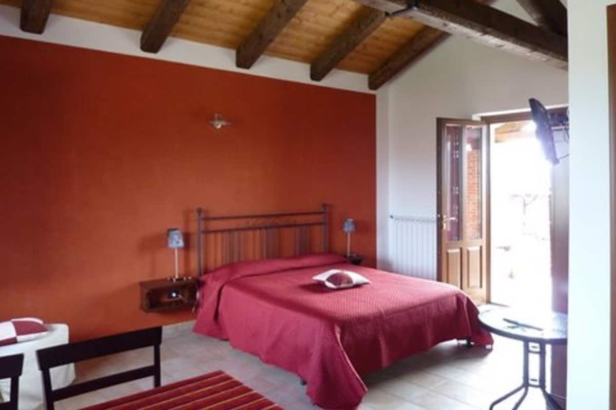 Une chambre avec un plafond en bois, un lit recouvert d’une couverture rouge, une table de chevet avec une lampe et une fenêtre donnant sur l’extérieur.