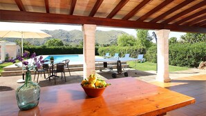 Une terrasse extérieure avec une table en bois, un saladier rempli de fruits et un vase de fleurs.