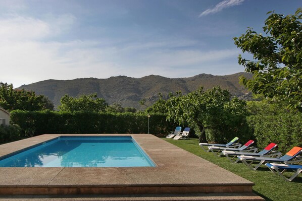 Une piscine rectangulaire à l’eau d’un bleu limpide, entourée d’une terrasse en béton. Plusieurs chaises longues avec des coussins colorés sont disposées autour de la piscine. Au fond, on aperçoit des arbres verts et une chaîne de montagnes sous un ciel dégagé.