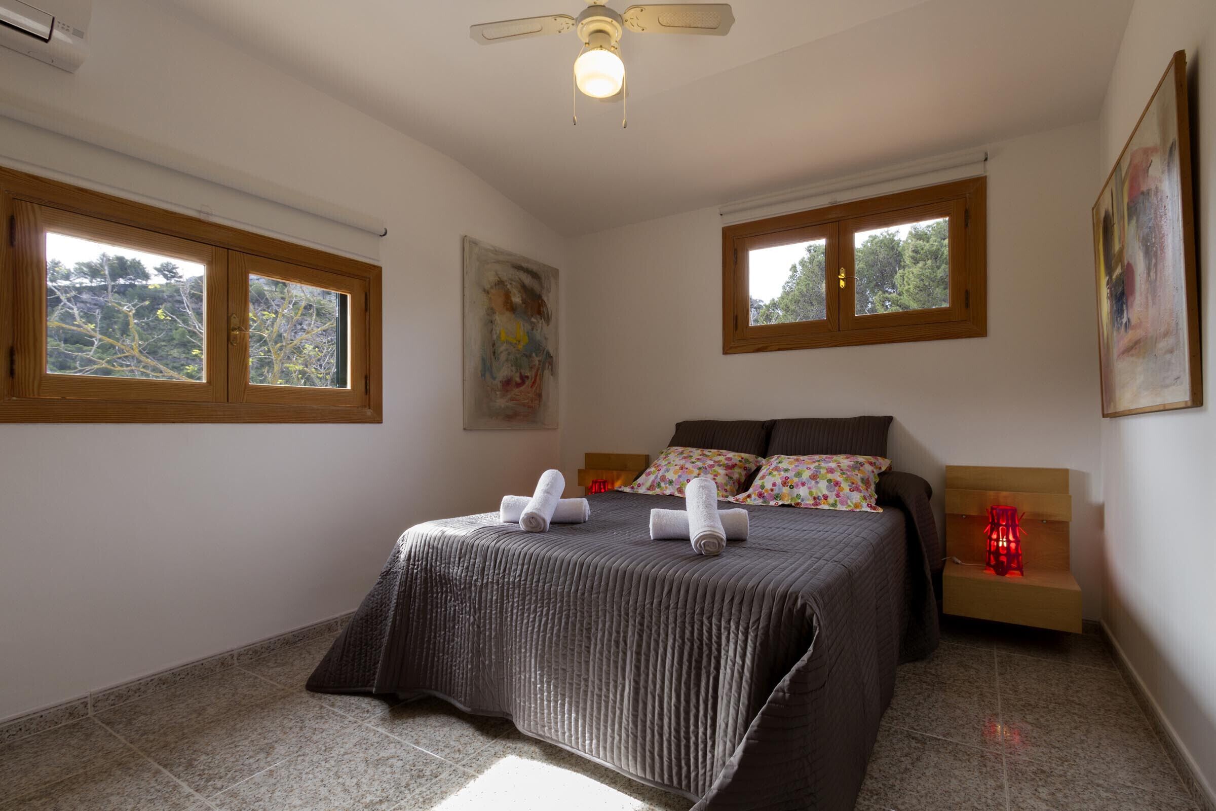 Ein Schlafzimmer mit einem Bett, zwei Fenstern, einem Deckenventilator und einem Nachttisch mit einer roten Lampe.