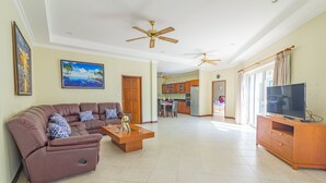 A spacious living room with a brown sofa set, a wooden coffee table, and a TV on a wooden stand. There are ceiling fans and a kitchen in the background. - 2 Bed Majestic Villa Near Beach (Pattaya)