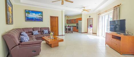 A spacious living room with a brown sofa set, a wooden coffee table, and a TV on a wooden stand. There are ceiling fans and a kitchen in the background.