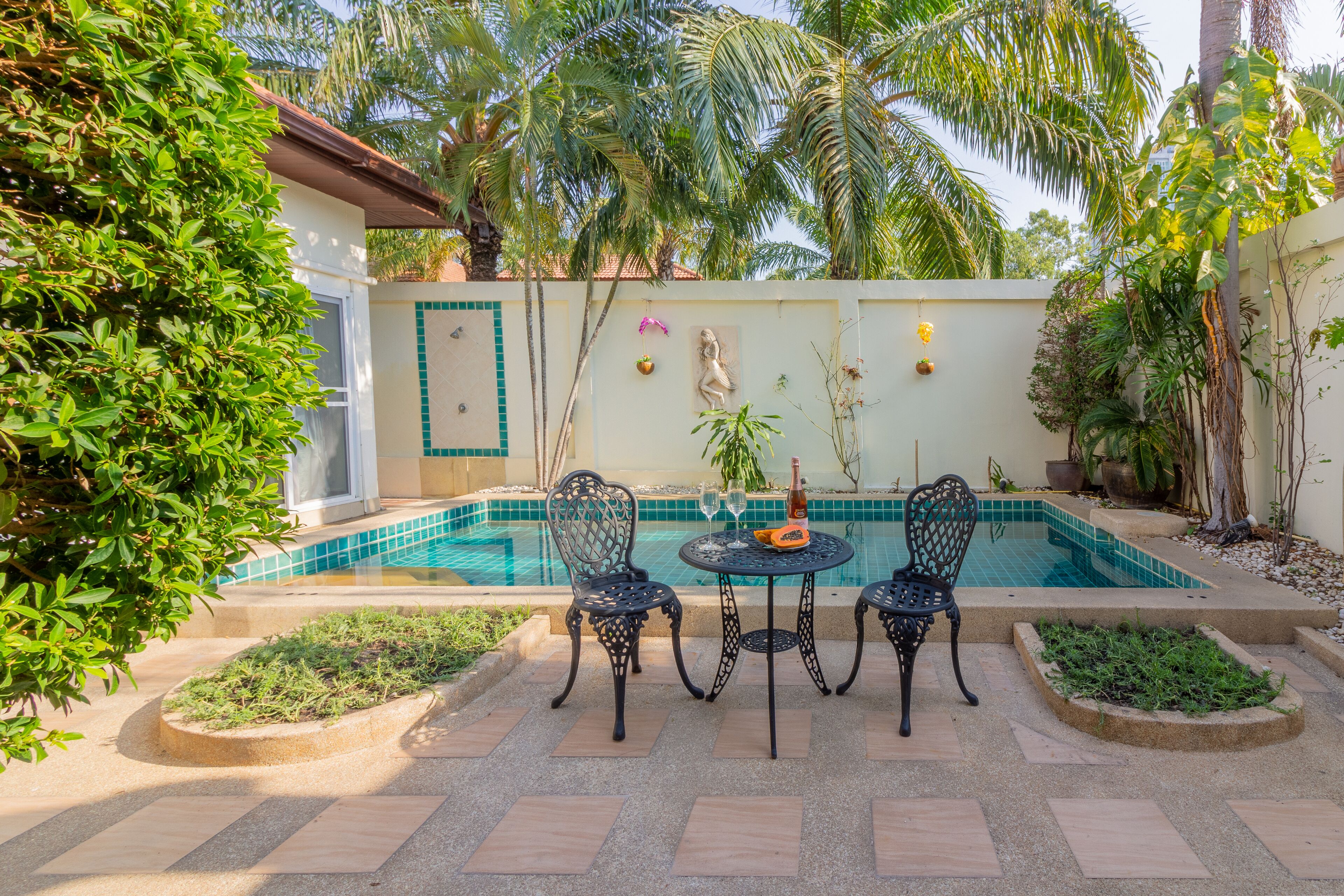 Uma área na beira da piscina com mesa e cadeiras, cercada por plantas e um painel de azulejos.