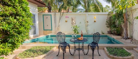 Uma área na beira da piscina com mesa e cadeiras, cercada por plantas e um painel de azulejos.