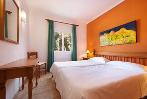 A hotel room with two beds, a wooden desk, a chair, a window with curtains, a painting on the wall, and a nightstand with a lamp.