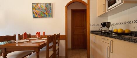 A dining area with a wooden table and chairs, a painting on the wall, and a kitchen with a microwave and lemons on the counter.