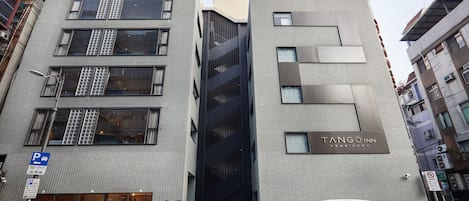 Il s’agit d’un bâtiment moderne, sur lequel on peut voir une enseigne indiquant « Tango Inn ». Il se distingue par son entrée imposante avec une façade en verre, de nombreuses fenêtres et un espace de stationnement où sont garées des voitures.