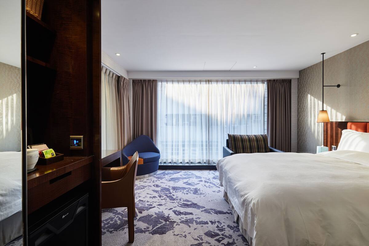 Une chambre d’hôtel avec deux lits, un bureau, une chaise et une grande fenêtre avec des rideaux.