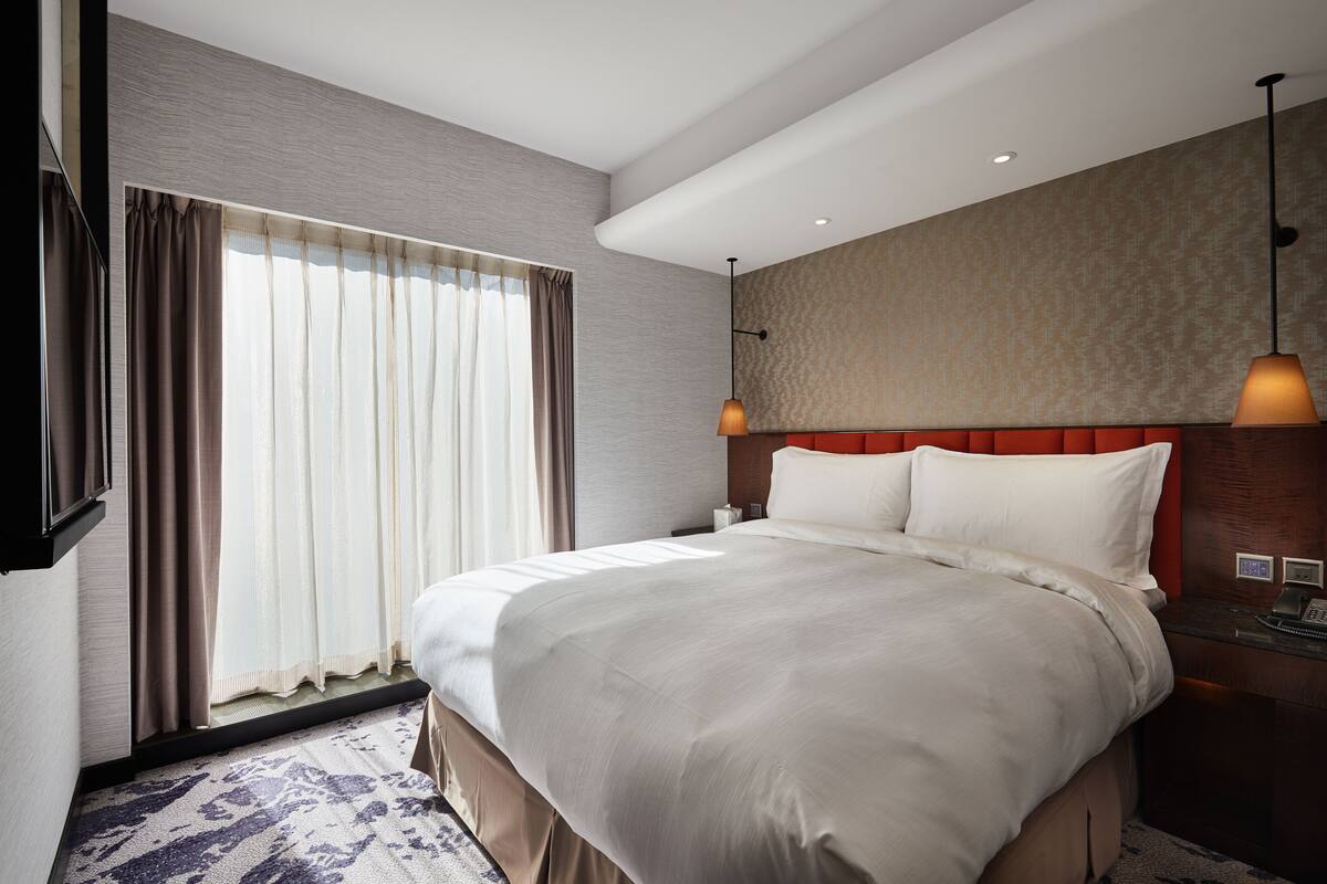 Une chambre d’hôtel moderne avec un grand lit, une tête de lit en bois, un bureau avec un téléphone et une grande fenêtre avec des rideaux.