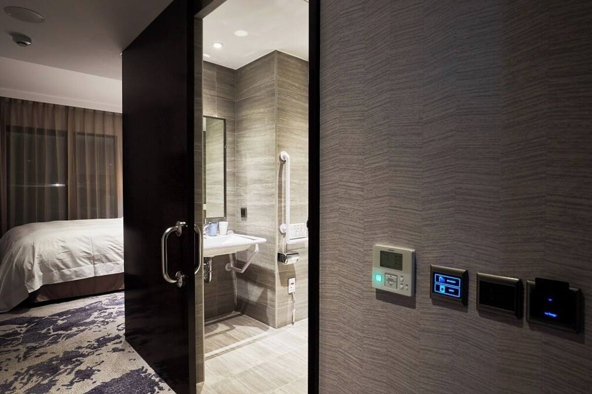Une chambre d’hôtel moderne équipée d’un lit, d’une salle de bain avec un lavabo et un miroir, et d’un panneau de commande mural.