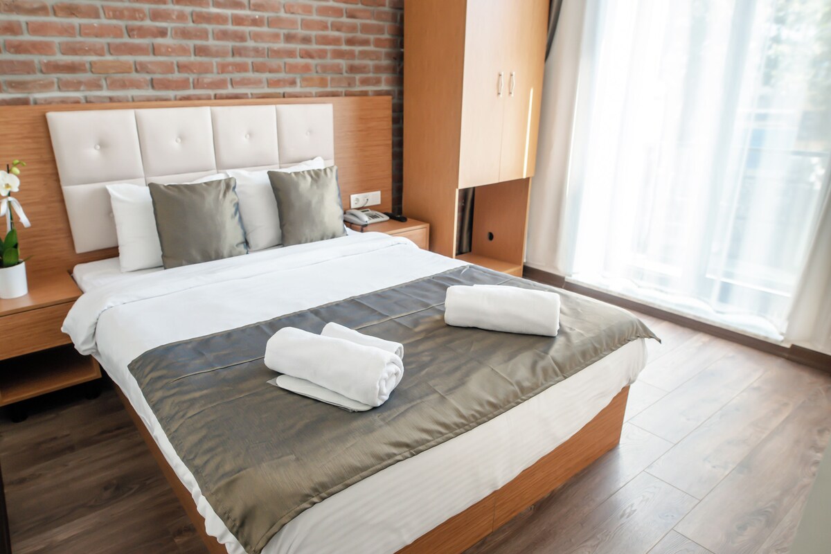 Una cama bien tendida con edredón gris y blanco, cabecera de madera y mesita de noche con lámpara y teléfono.