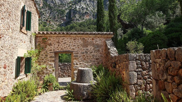 Un edificio de piedra con una entrada rústica, rodeado de follaje y montañas al fondo.