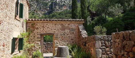A stone building with a rustic entrance, surrounded by greenery and mountains in the background.