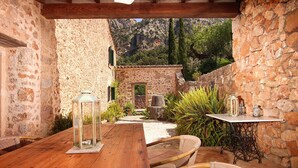Un patio al aire libre con una mesa de madera, sillas de mimbre, una pared de piedra y vistas a un paisaje montañoso.