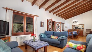 Un salon avec un canapé, une table basse et un téléviseur. Il y a une fenêtre avec des rideaux, un radiateur et un plafond en bois.